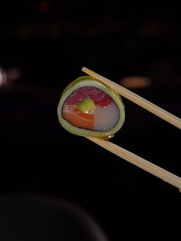 Vibrant sushi roll held by chopsticks against a dark background, showcasing fresh ingredients.