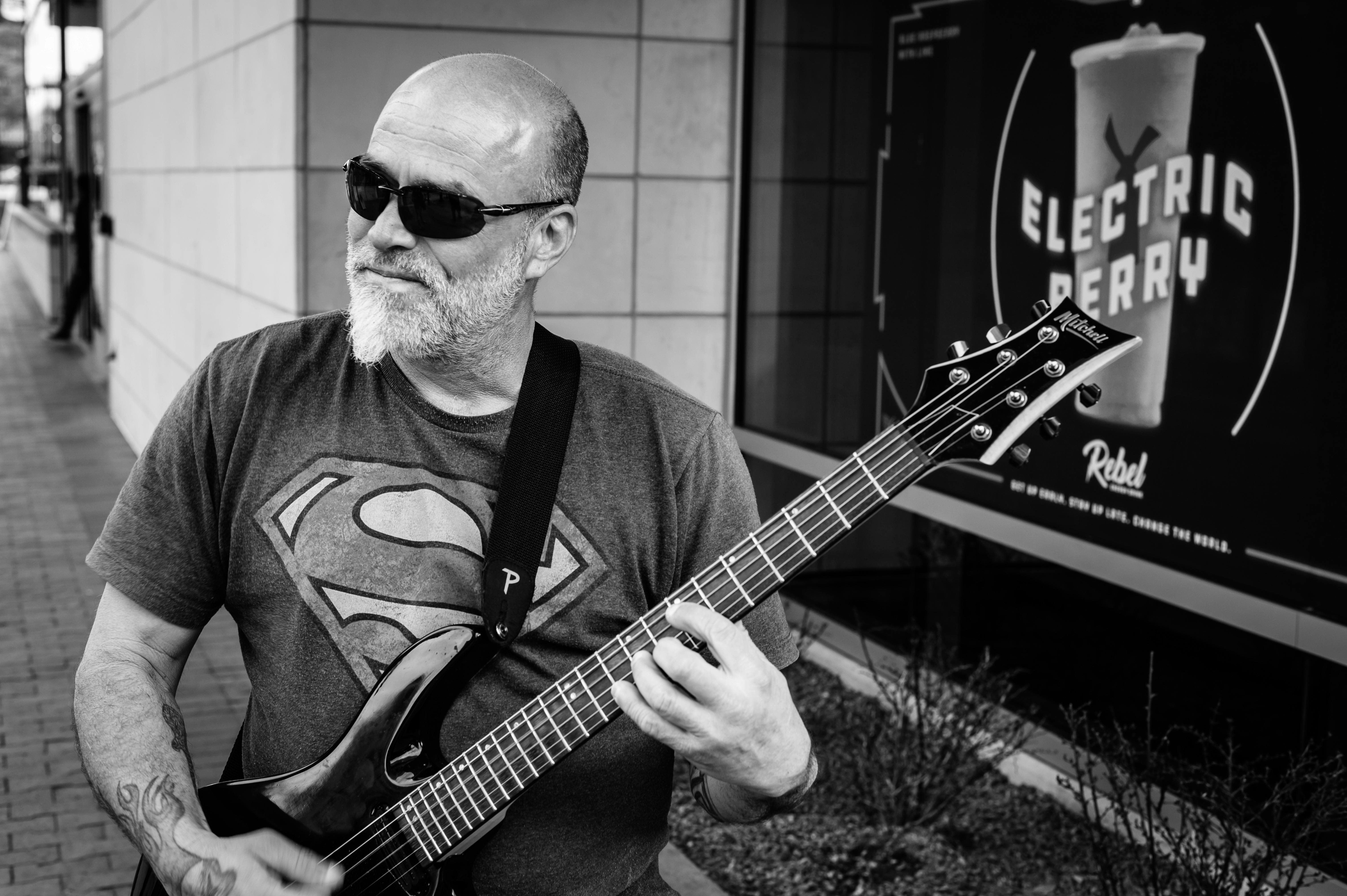 A bearded street musician plays an electric guitar, wearing shades and a Superman shirt.