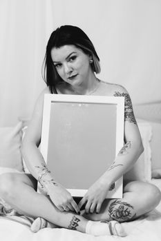 Artistic portrait of a tattooed woman sitting with a blank frame, captured in black and white.