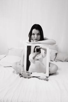 Artistic black and white photo of a woman sitting on a bed holding a mirror showing her reflection.