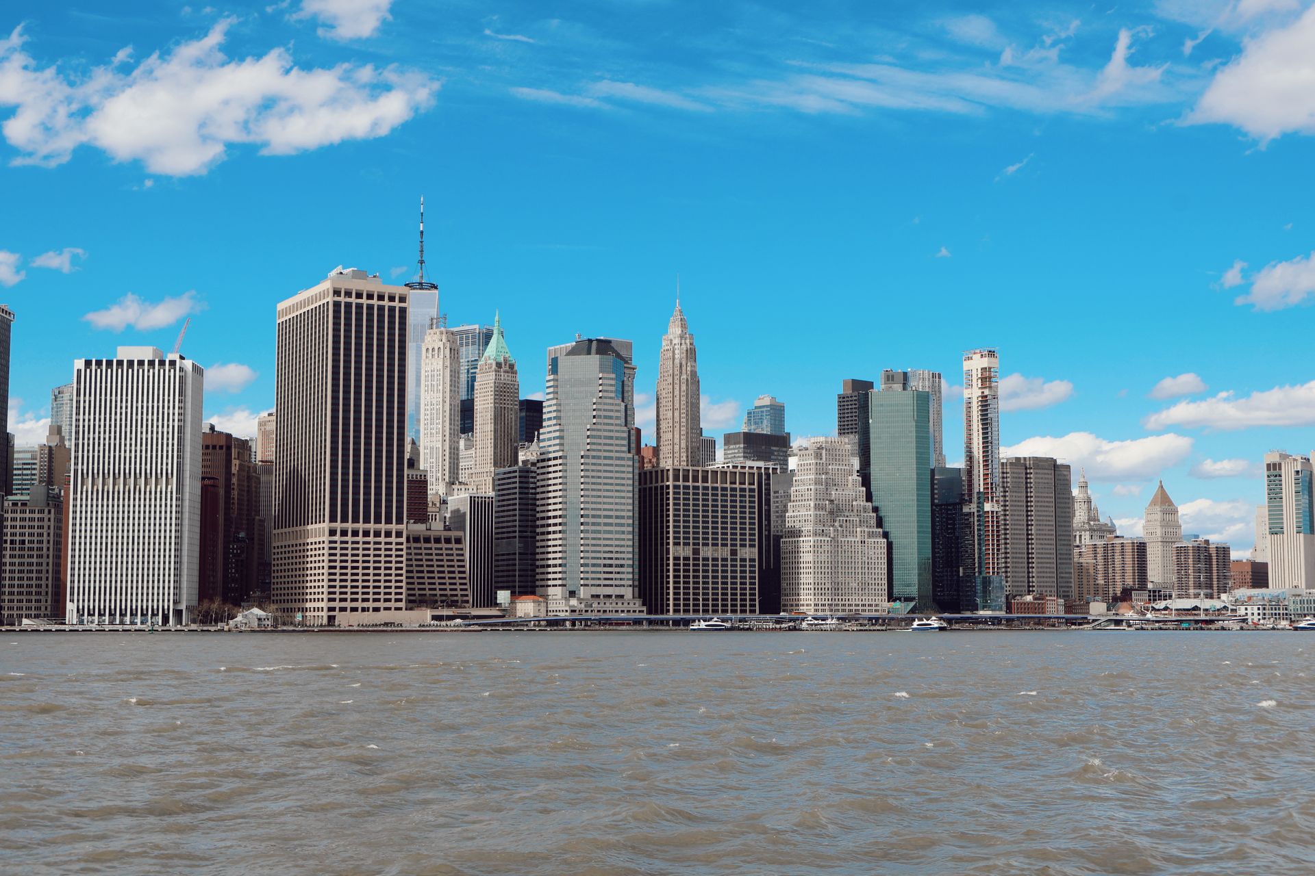 Bright daytime view of Manhattan skyline with iconic skyscrapers reflected in the East River under clear blue sky
