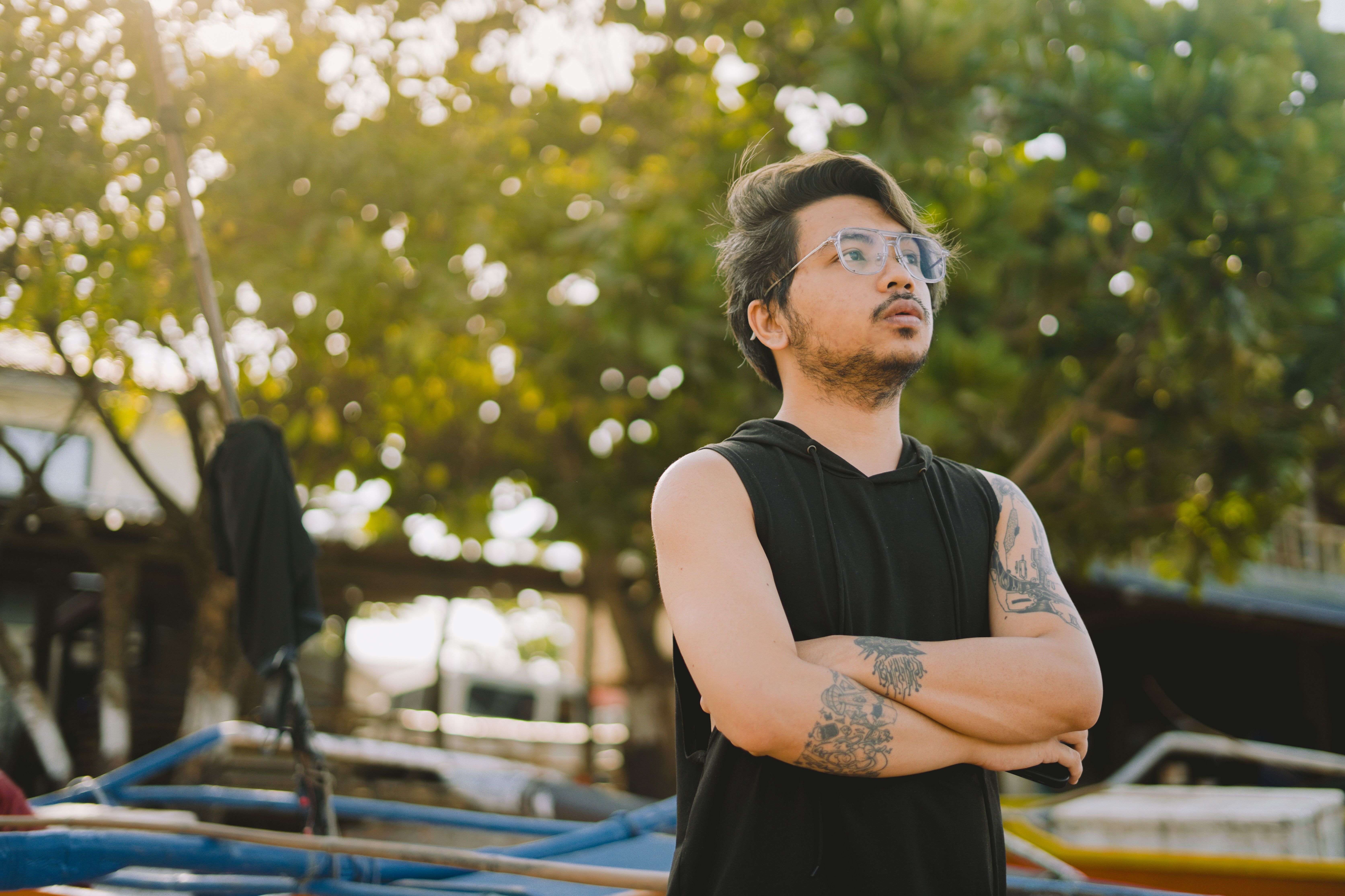A man with tattoos and eyeglasses stands outdoors, looking thoughtful in a casual sleeveless top.