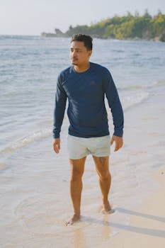Man in blue sweatshirt walking barefoot on a sunny beach. Casual summer vibe.