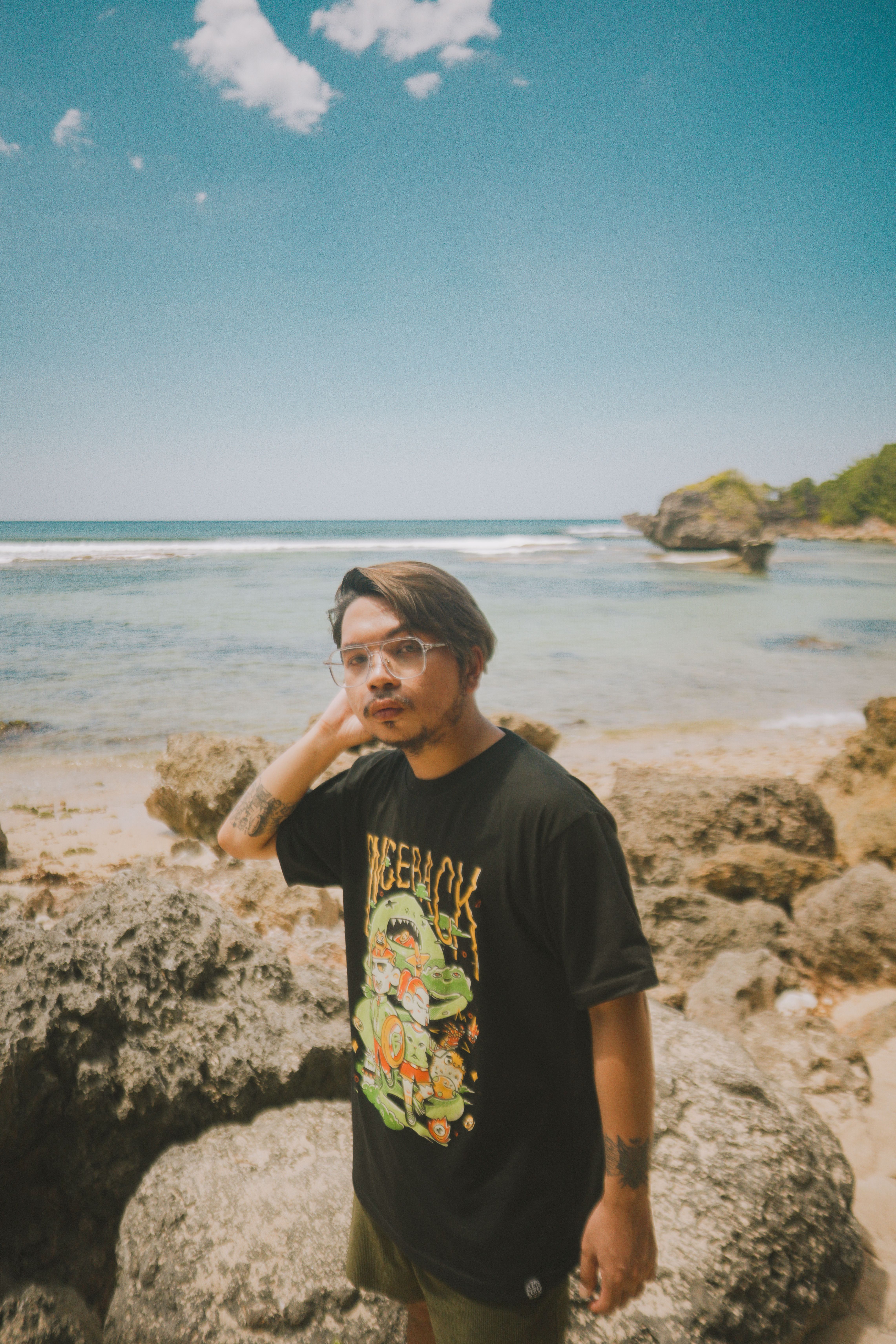 Stylish young man in black T-shirt posing by the rocky seashore in Bolinao, Philippines under a bright blue sky.