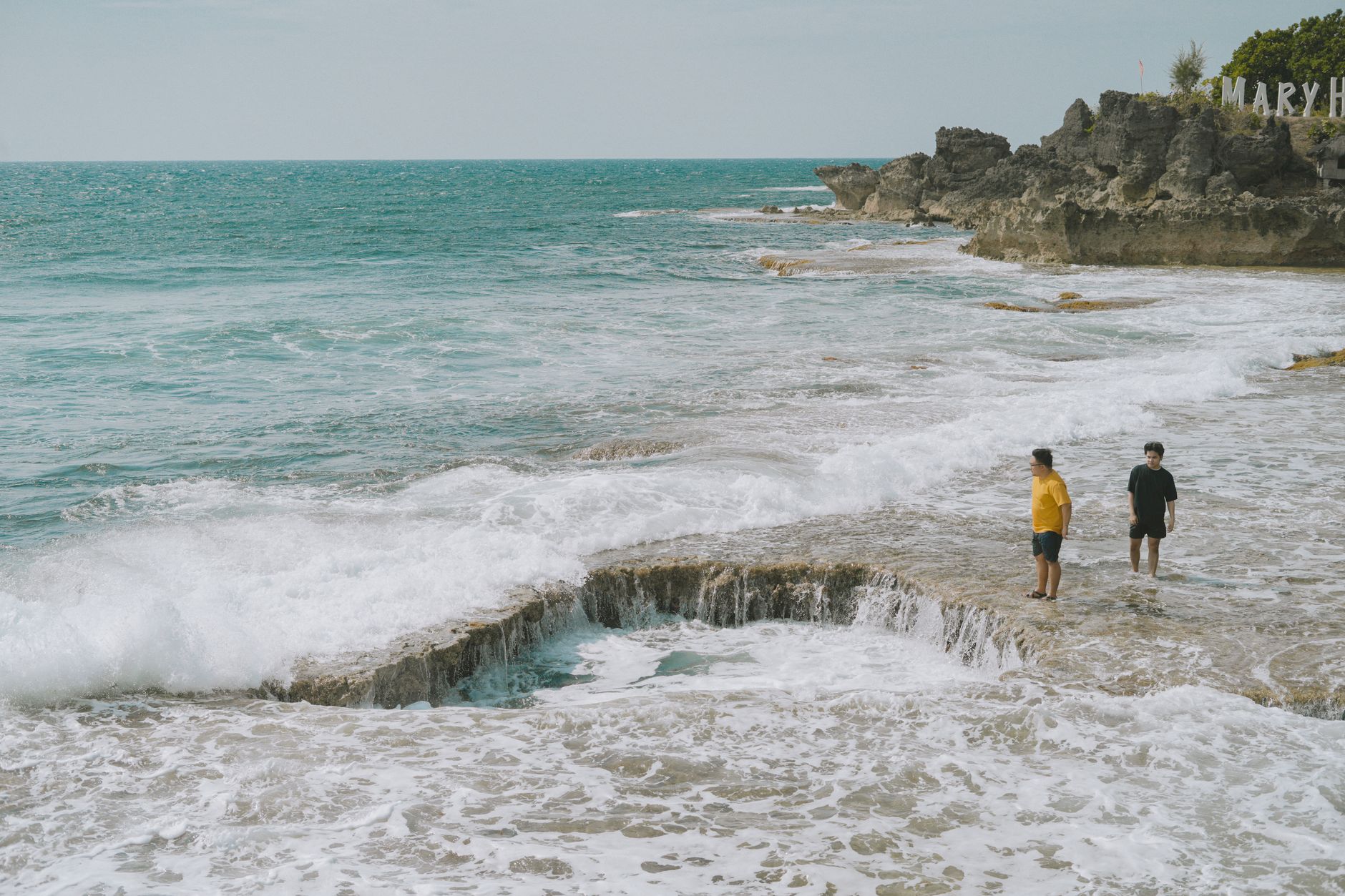 Two men stand on the rocky shore experiencing the waves in Bolinao, Philippines, on a summer day.