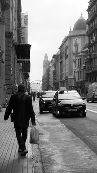 Black and white city street with pedestrian walking along sidewalk, capturing urban life.