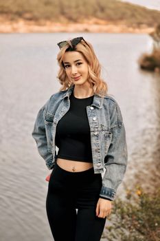 A young woman in a denim jacket posing by a tranquil lake on a sunny day.