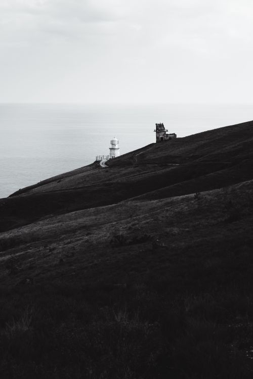 Free Dramatic black and white landscape featuring a lighthouse on a coastal hill under a cloudy sky. Stock Photo