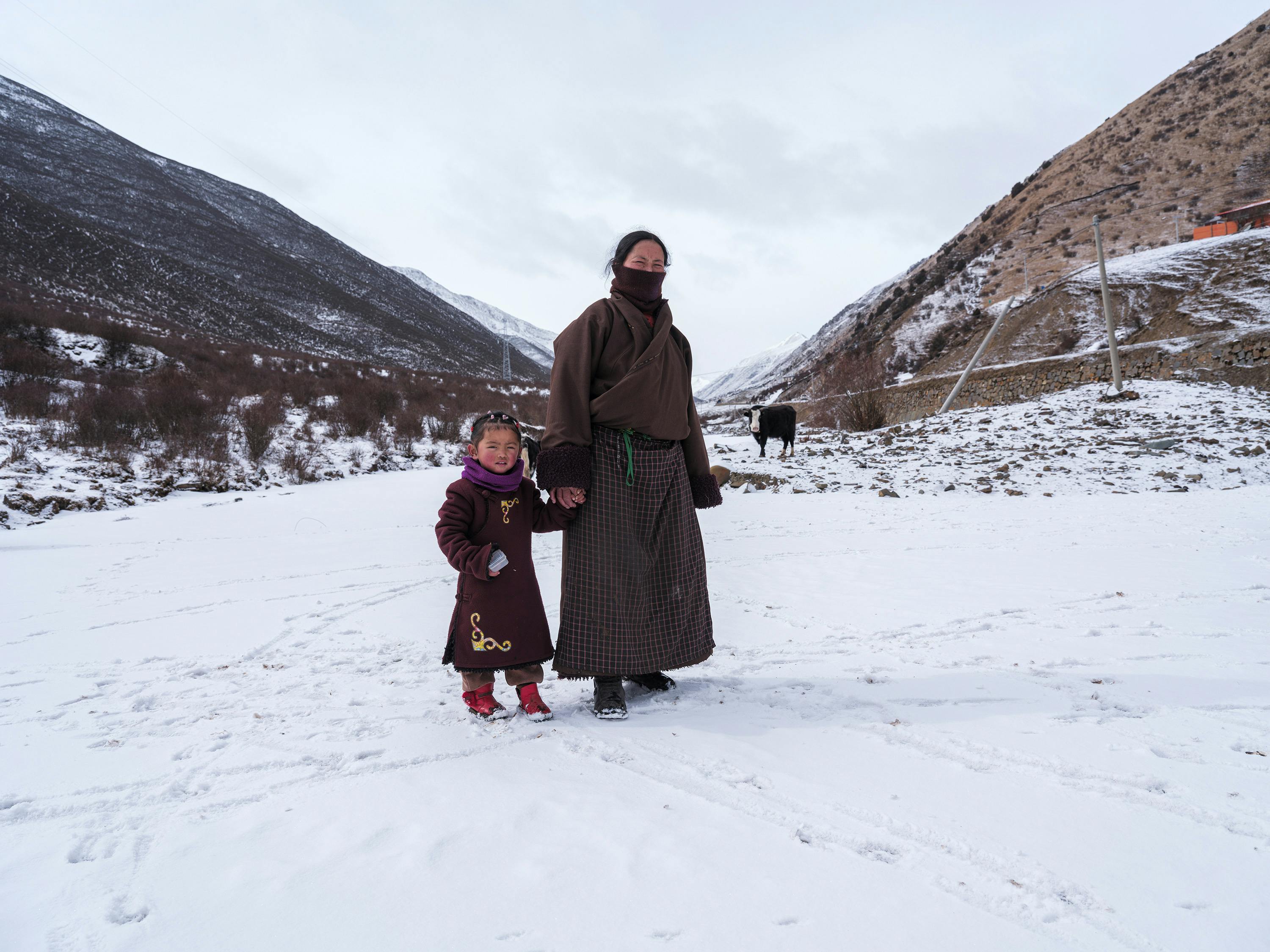 A mother and daughter in traditional clothing stand on a snowy landscape in a rural winter setting.