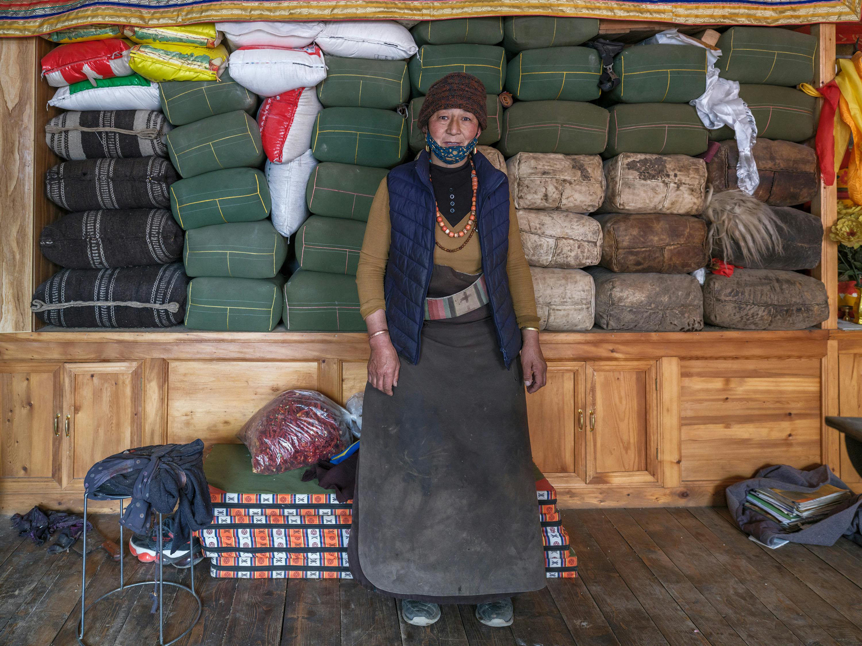 Asian woman standing indoors with winter attire and traditional clothing, surrounded by packed goods.