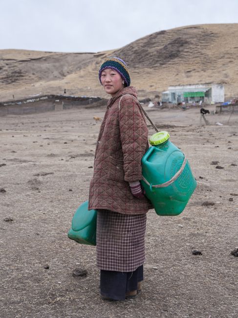 A Mongolian woman in a rural area carrying large green water containers, set against a barren desert landscape.