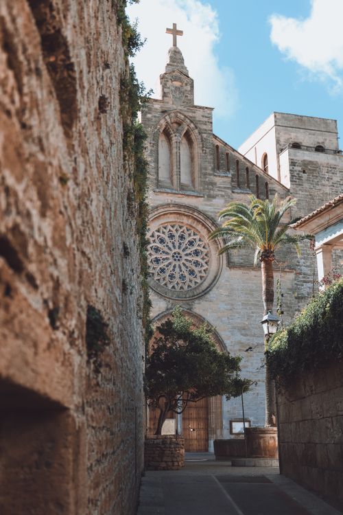 Free Narrow alley in Alcúdia leading to a classic stone church with intricate architectural details. Stock Photo