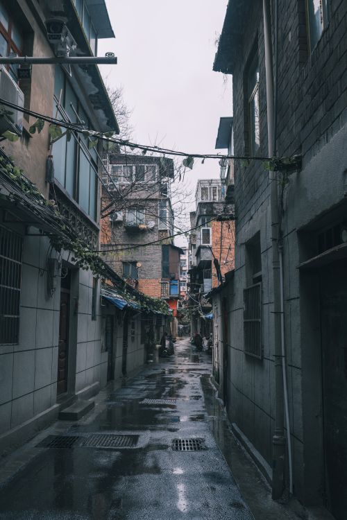 Free Moody urban alleyway with wet pavement reflecting city buildings on a cloudy day. Stock Photo