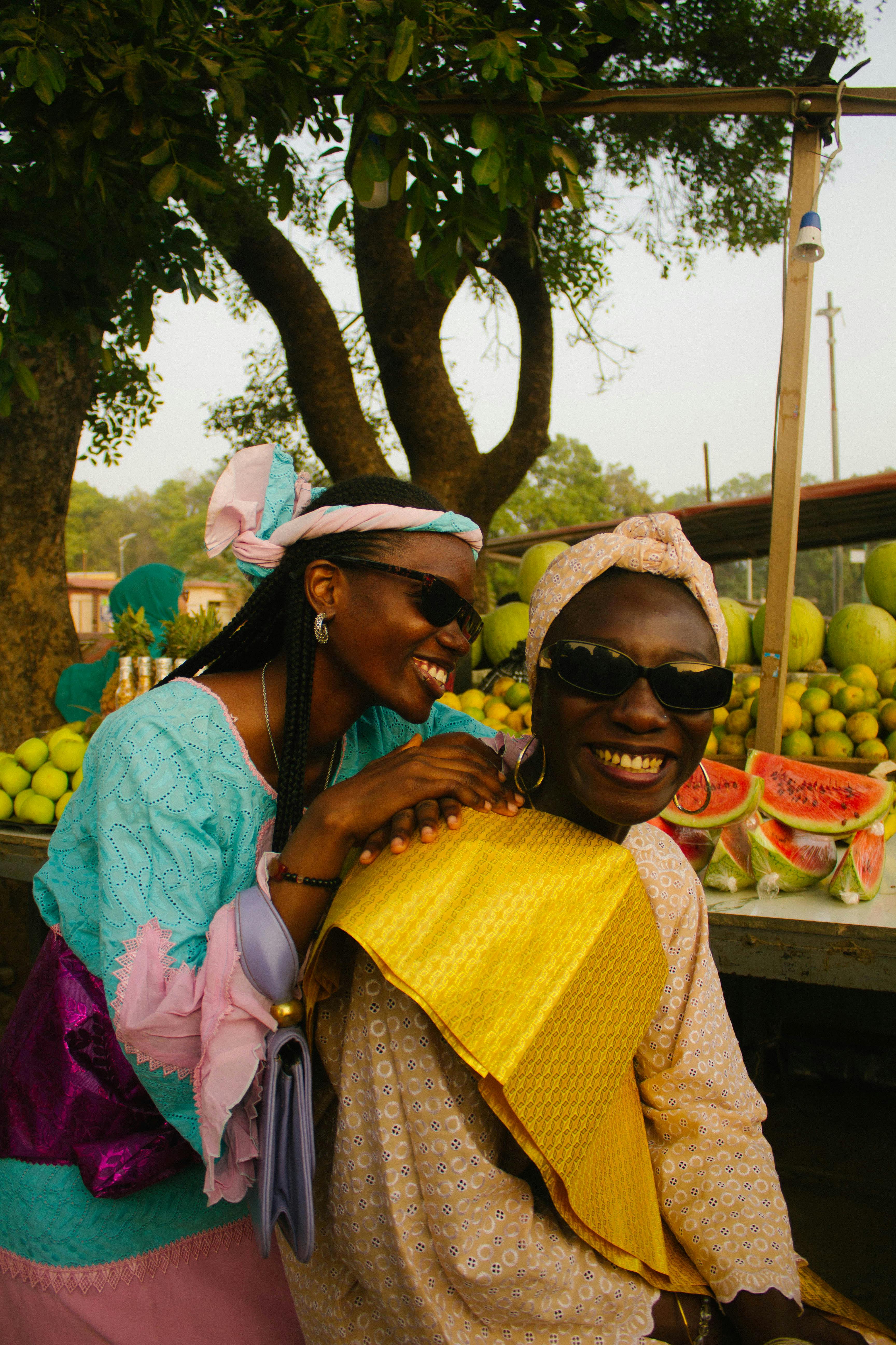 Two African women in vibrant traditional clothing enjoying a laugh at a lively fruit market.