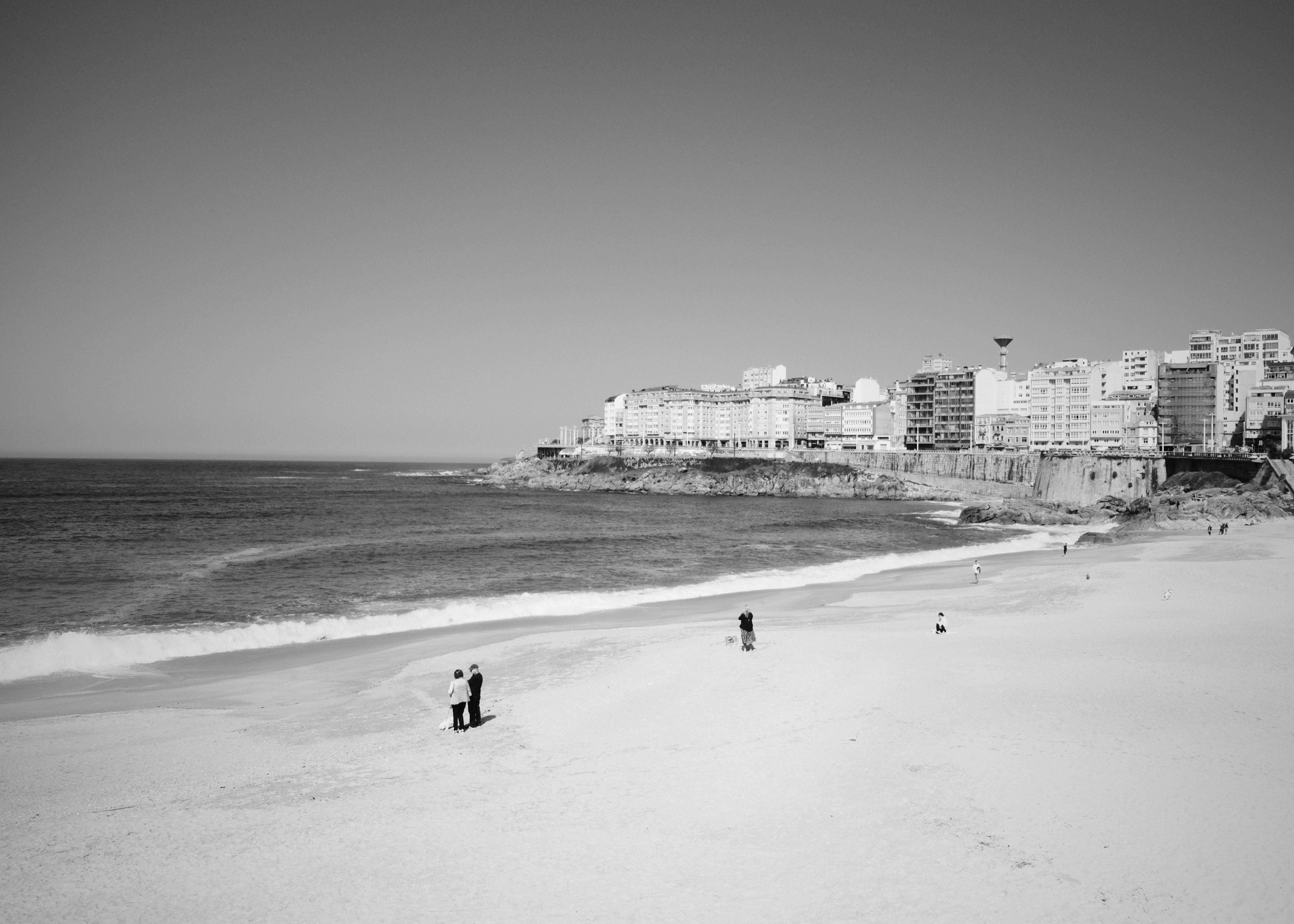 Black and white photograph of A Coruña beach with city skyline and ocean waves, capturing serene coastal life.