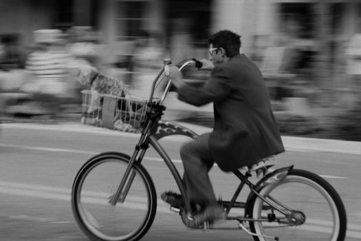 Black and white photo of a man cycling with a dog in the basket on a city street in motion.