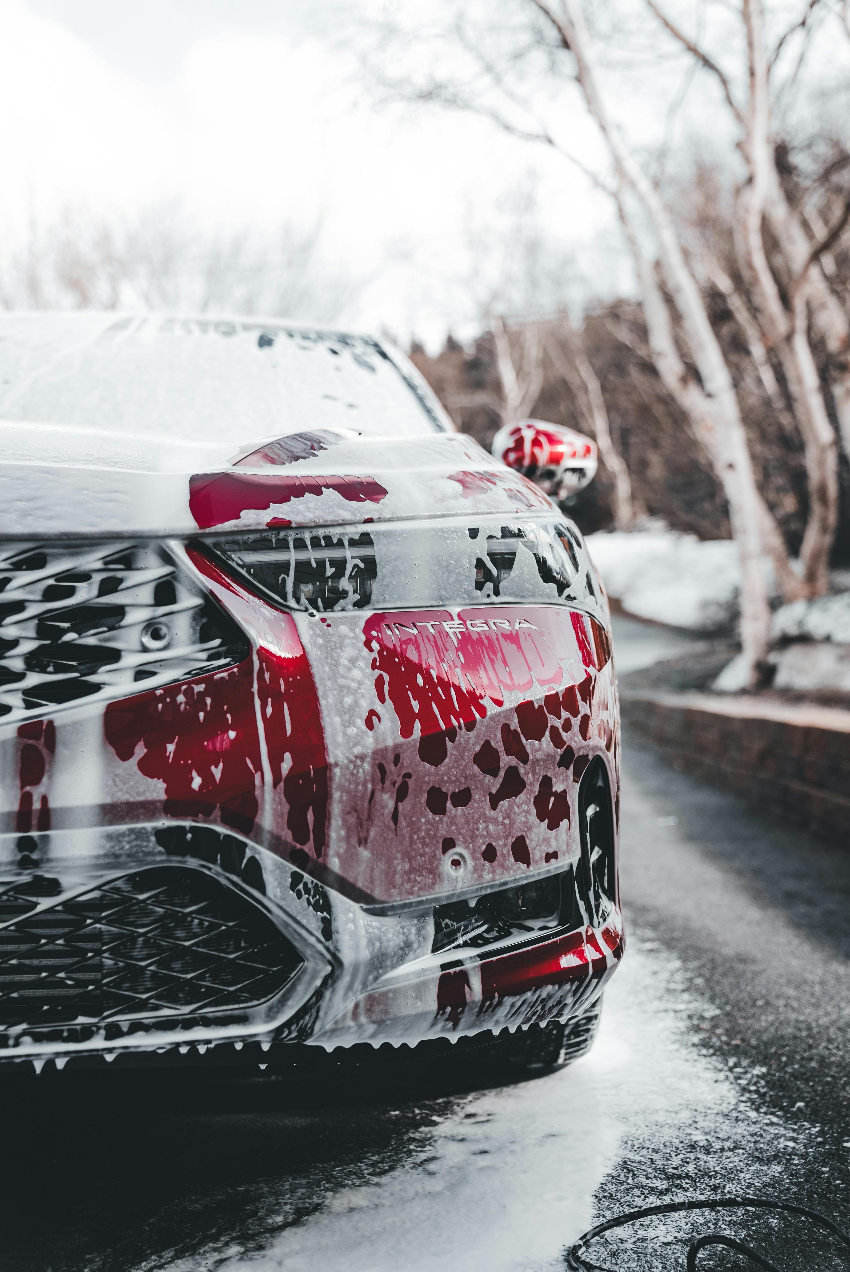Close-up view of a red car covered in foam during a car wash. The image showcases soap suds on the grille and headlight