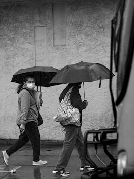 Black and white photo of people walking in the rain with umbrellas in a city setting.