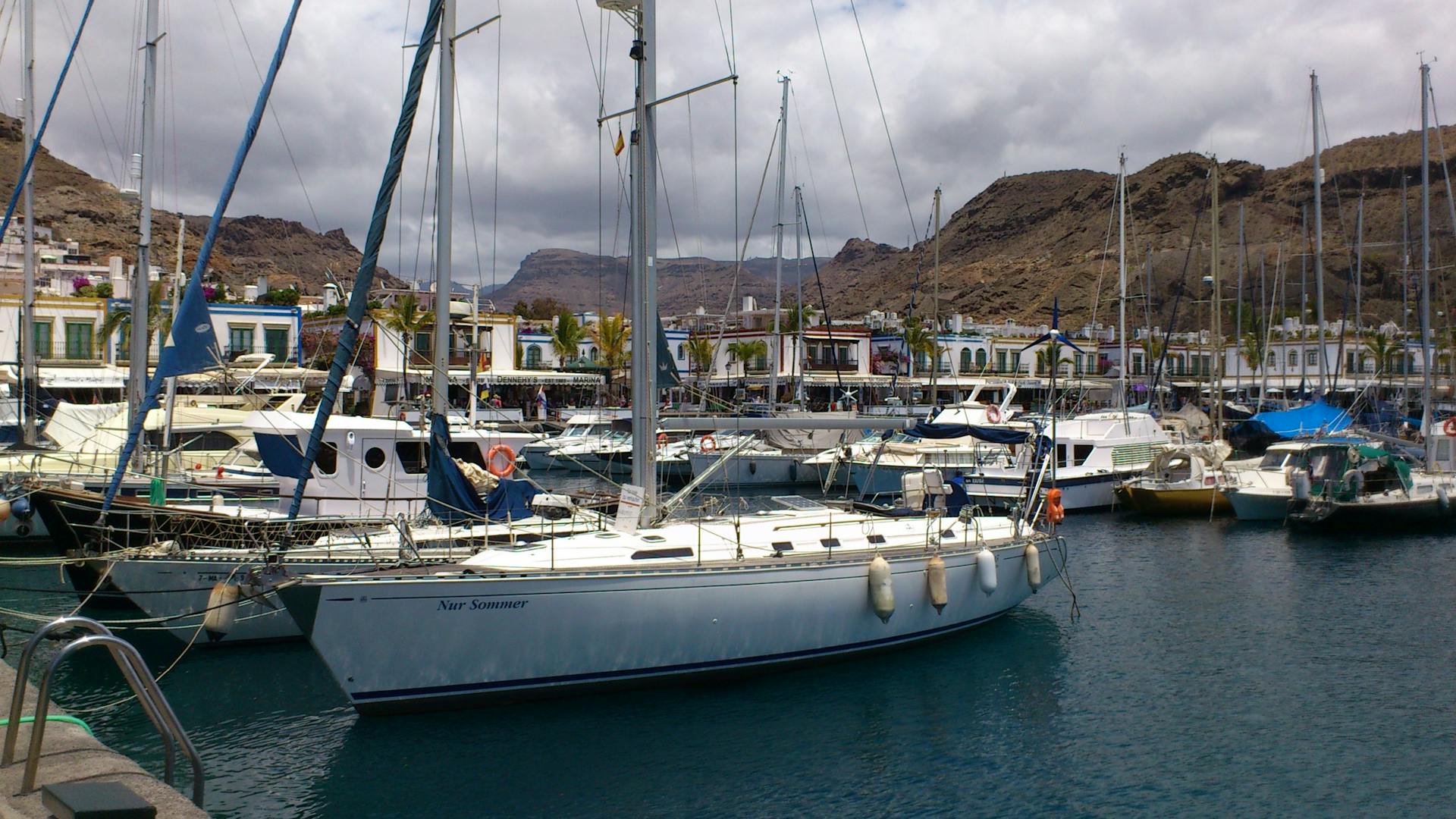 Yachts anchored in a marina under cloudy skies with mountains in the background.
