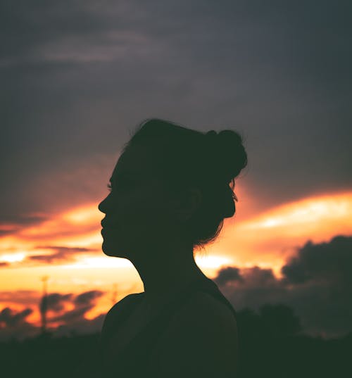 Side view of unrecognizable calm female with bunch hairstyle relaxing on street and admiring picturesque sunset sky