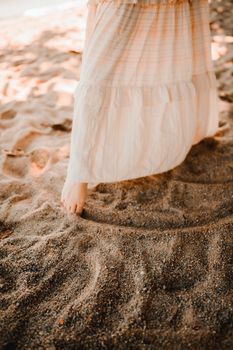 Close-up of woman's feet in a flowing dress on a sunny beach, highlighting summer vibes.