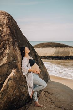 A serene pregnant woman in a white lace cover-up leans against a rock on a sandy beach.