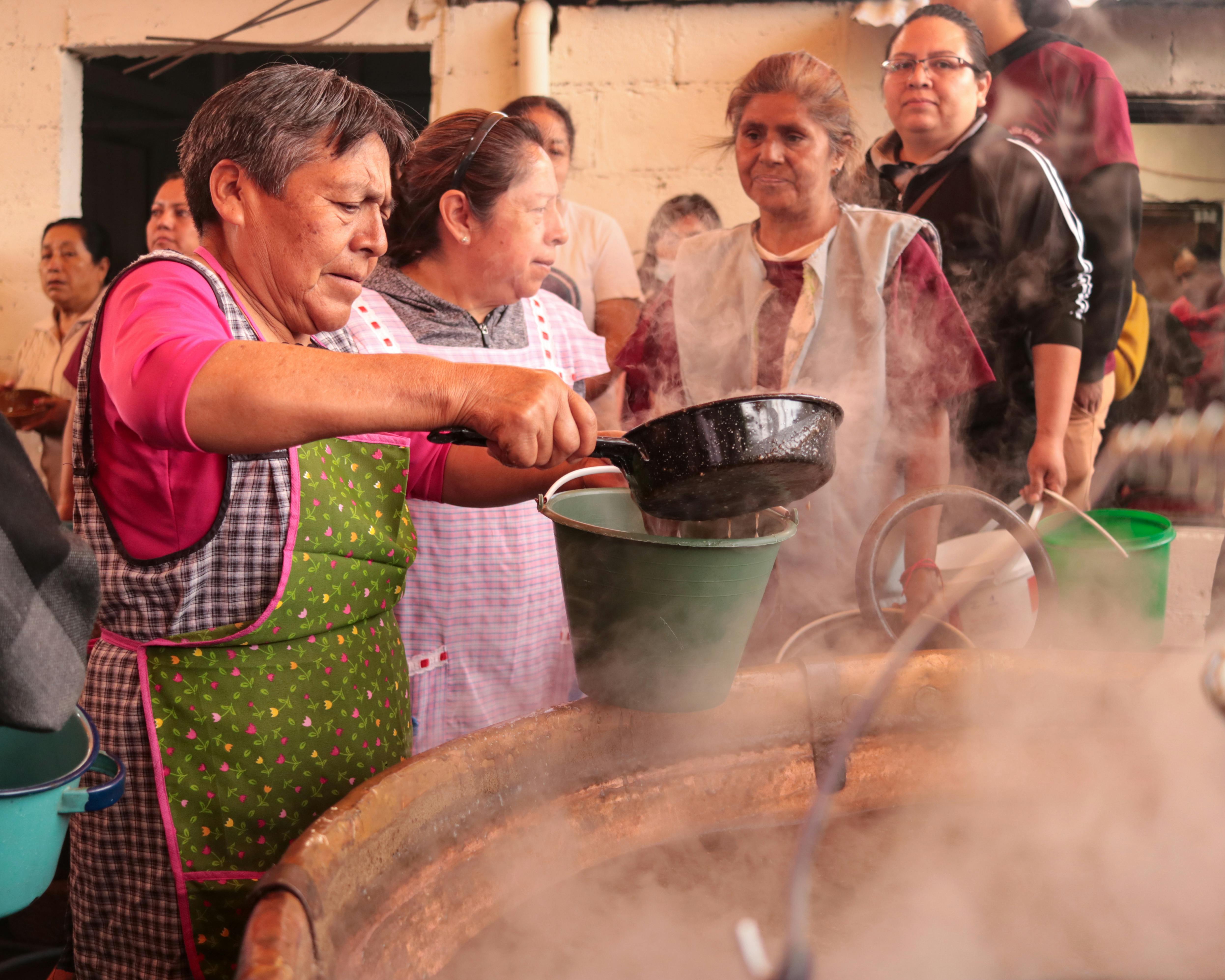 Close-up shot of a group of women in matching aprons serving steaming bowls of soup and fufu to a long line of people outside a modern church building
