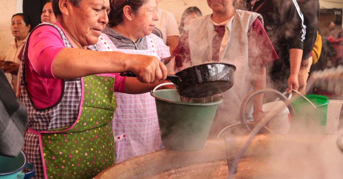 Close-up shot of a group of women in matching aprons serving steaming bowls of soup and fufu to a long line of people outside a modern church building