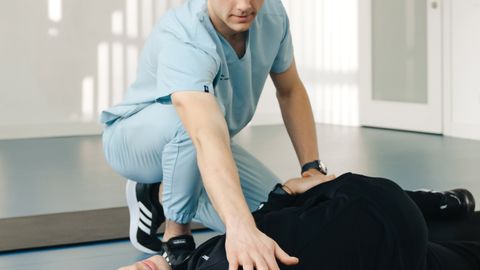 A physiotherapist assists an adult male patient in a stretching exercise on the floor indoors.
