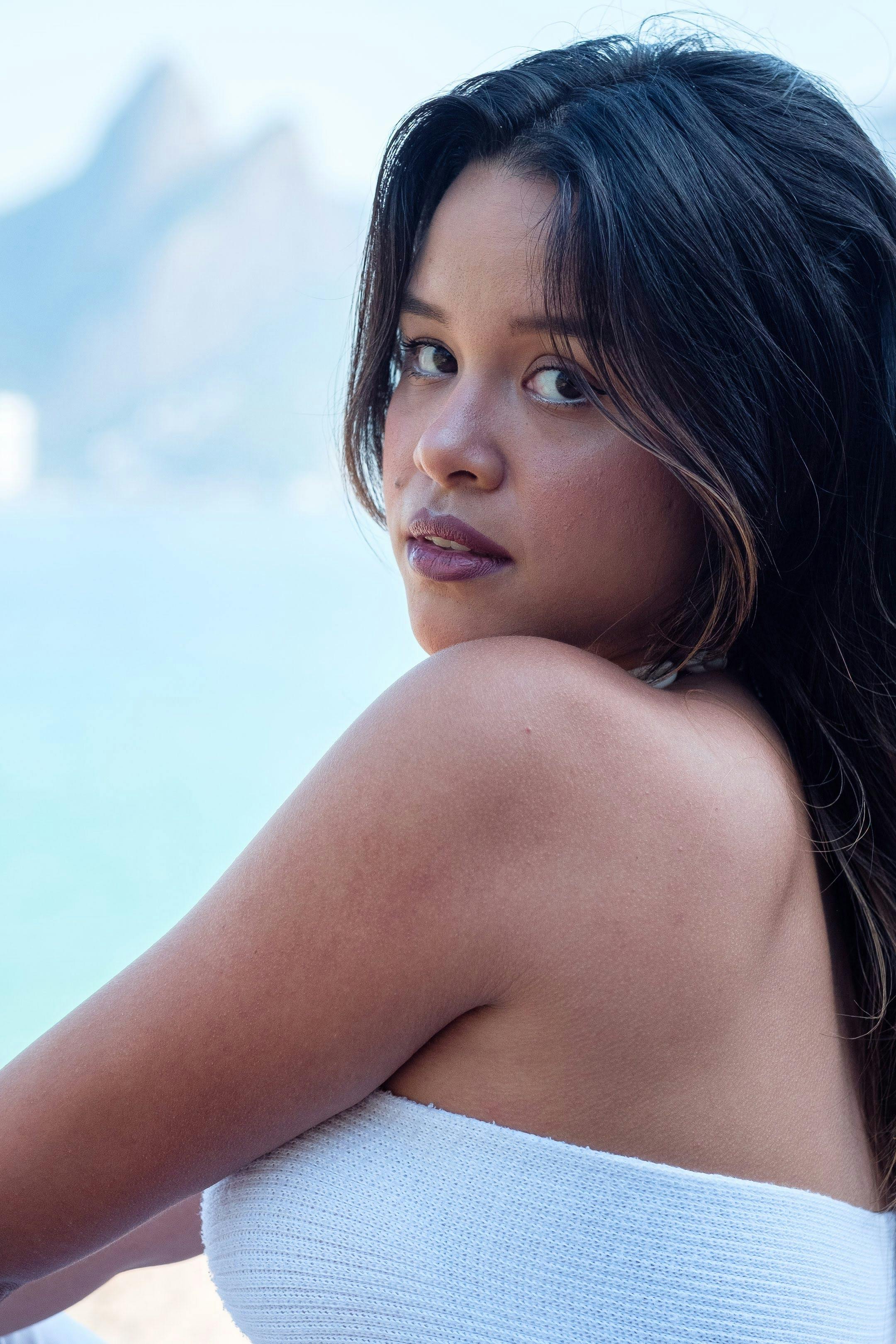 A fashionable woman in a white dress poses by a stunning coastline, featuring mountains.