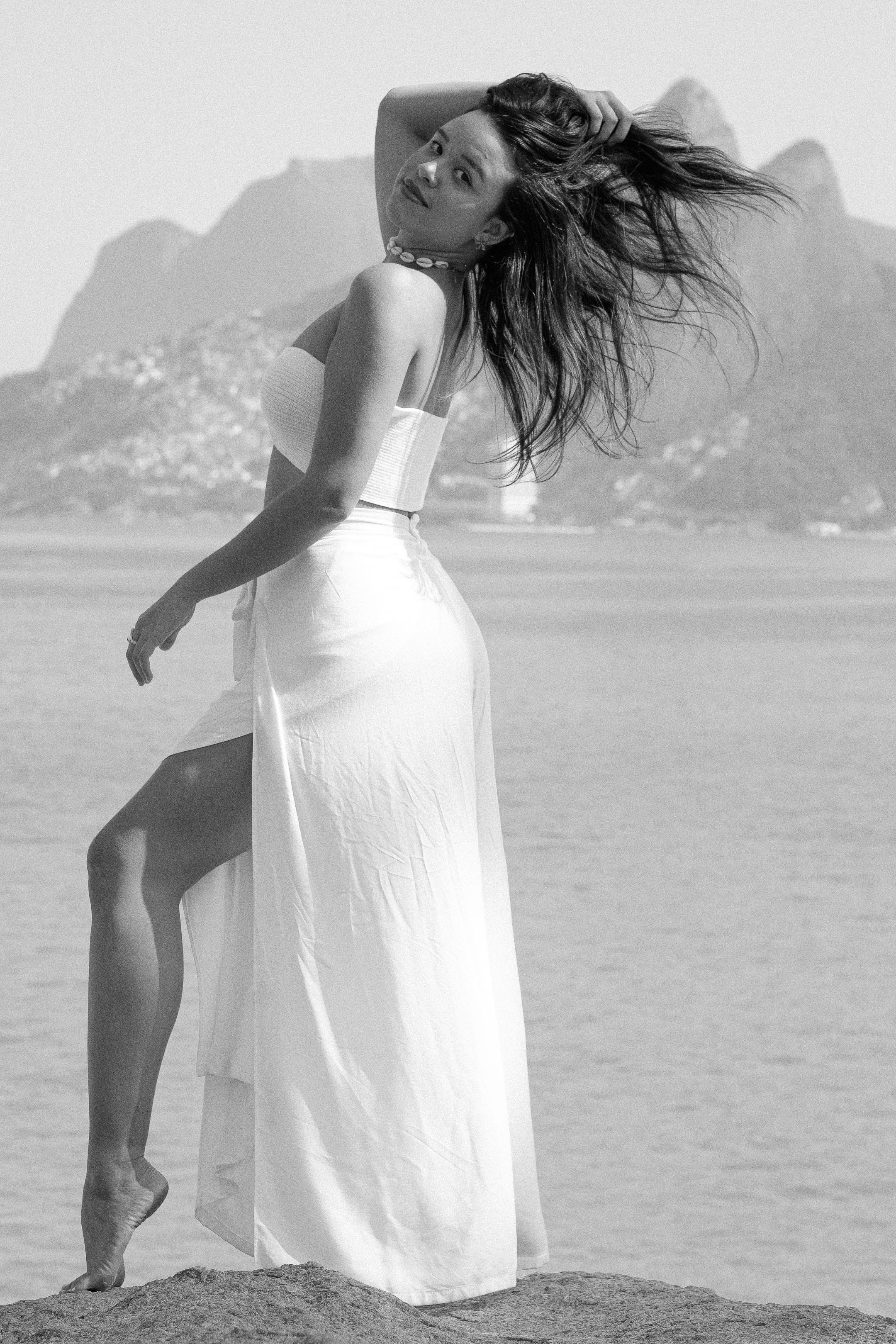 Graceful woman in a white dress striking a pose on a beach with a scenic backdrop.