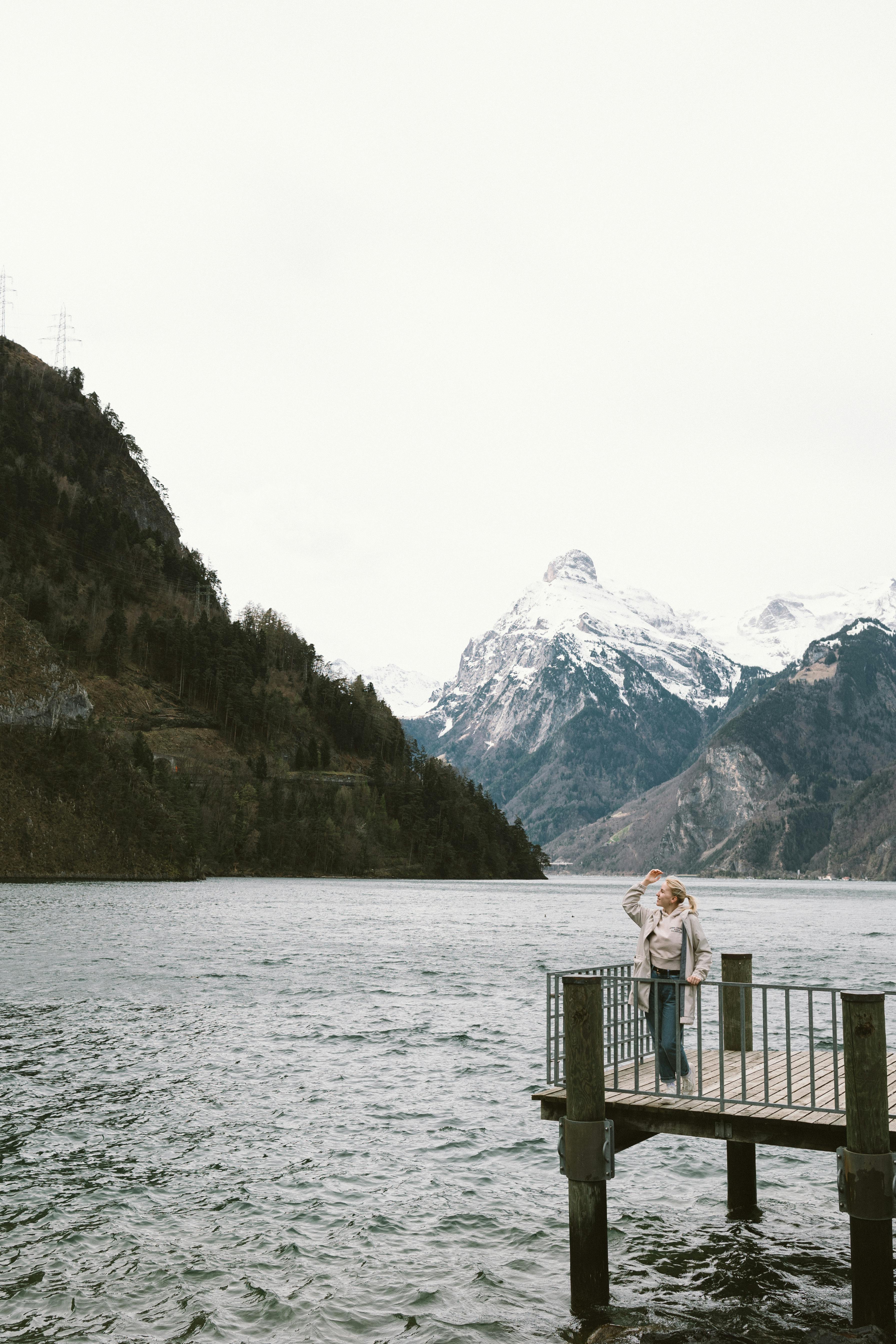 Woman standing on a pier by a tranquil lake with snow-capped mountains in the background.