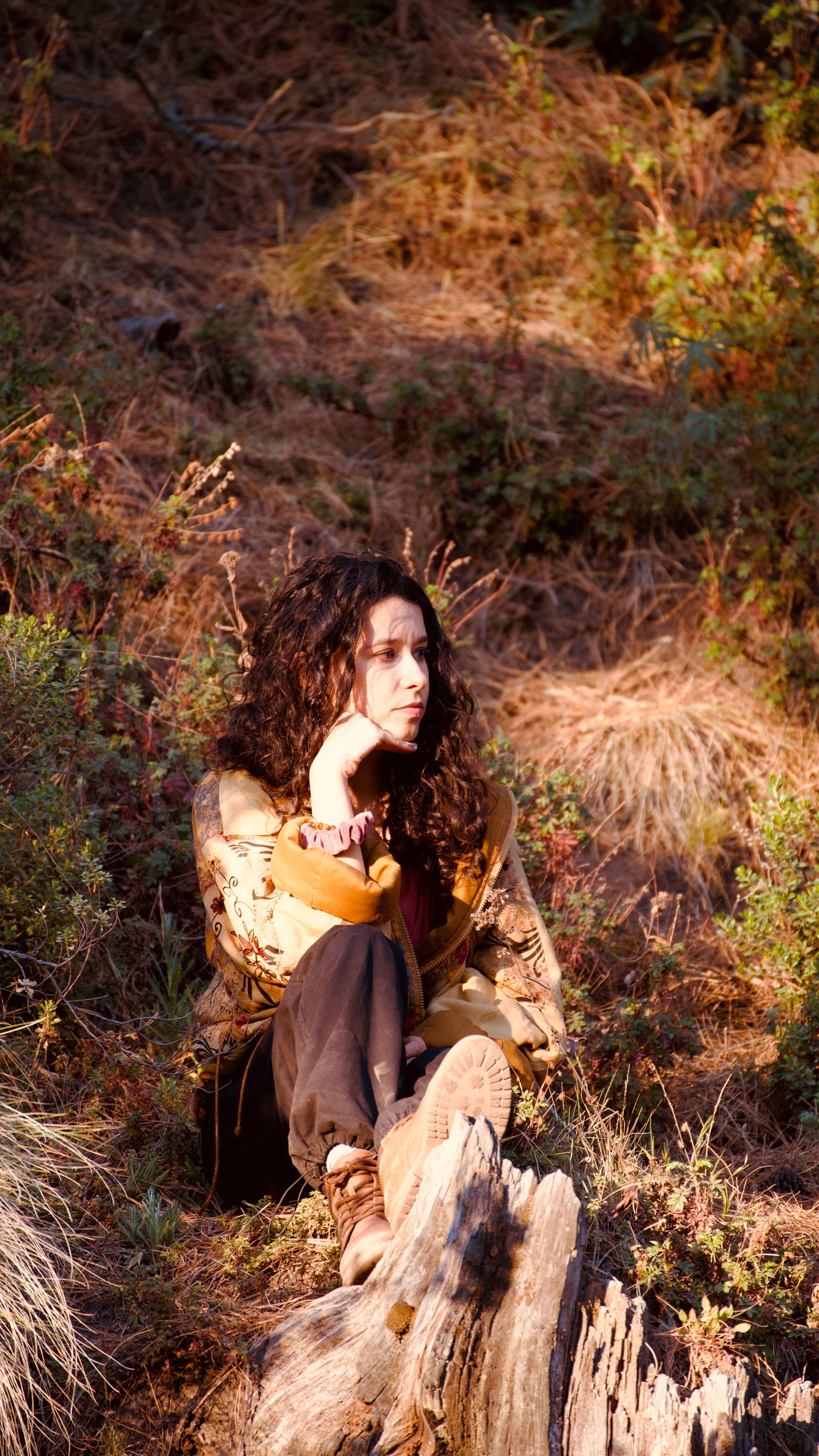 Woman with curly hair sitting outdoors in a sunny, rural meadow, embracing nature.