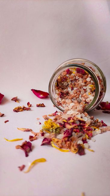 Fotomural Un tarro de vidrio de sales de baño aromáticas mezcladas con flores secas y hierbas derramado sobre un fondo blanco.