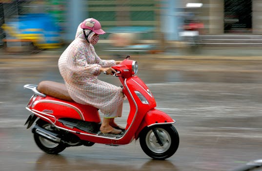 Action shot of a woman riding a red scooter on a rainy city street, wearing protective gear.