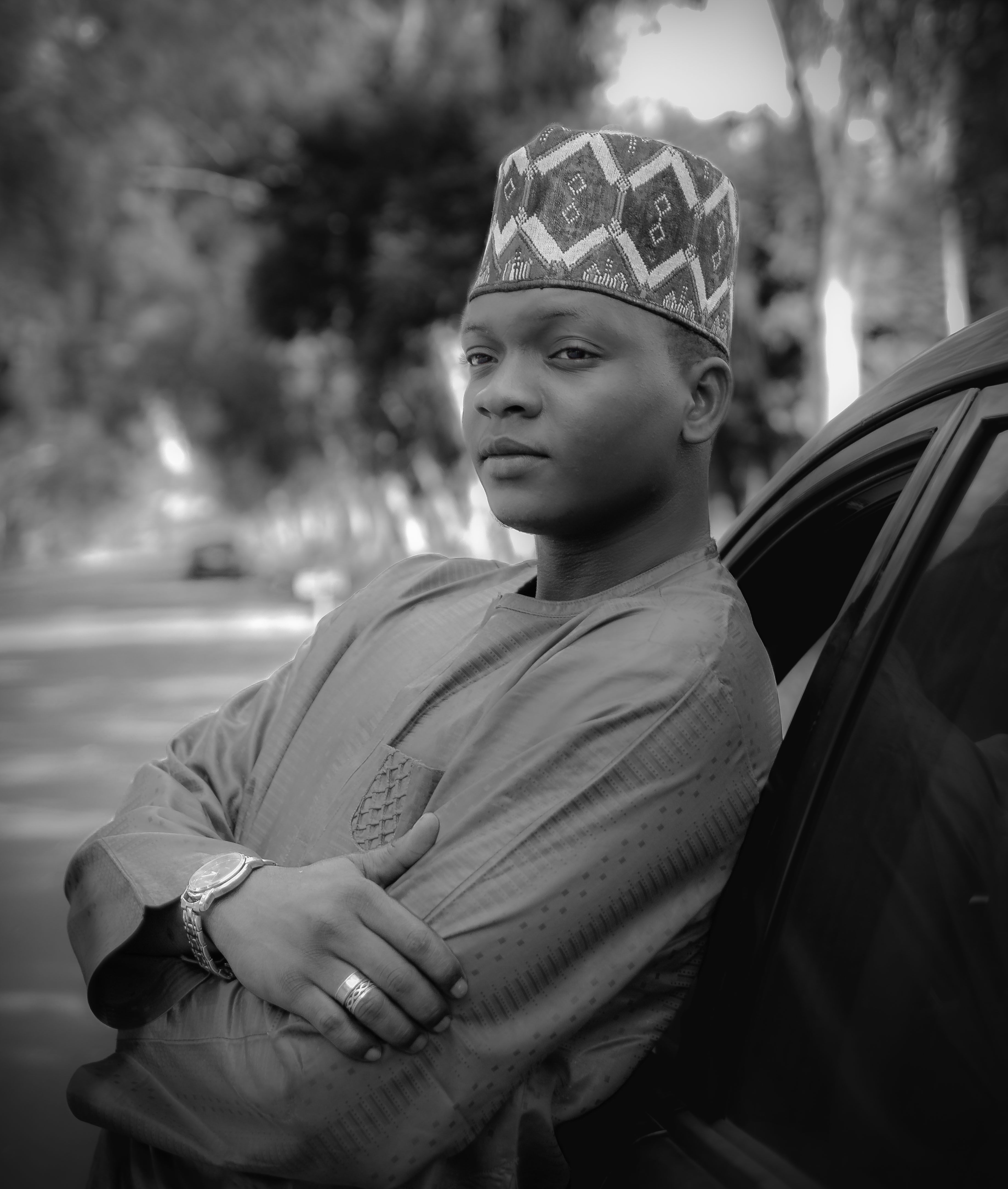 Black and white portrait of a man in traditional attire, leaning on a car outdoors.