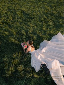 A bride in a white gown lies on grass, holding a bouquet, at sunset.