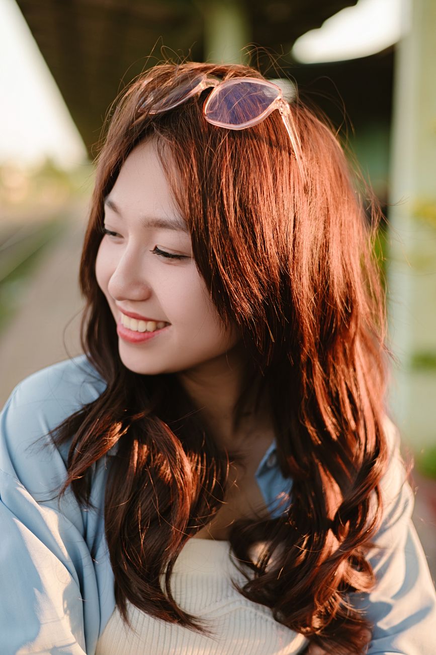 A young woman with eyeglasses on her head smiles outdoors, exuding happiness.