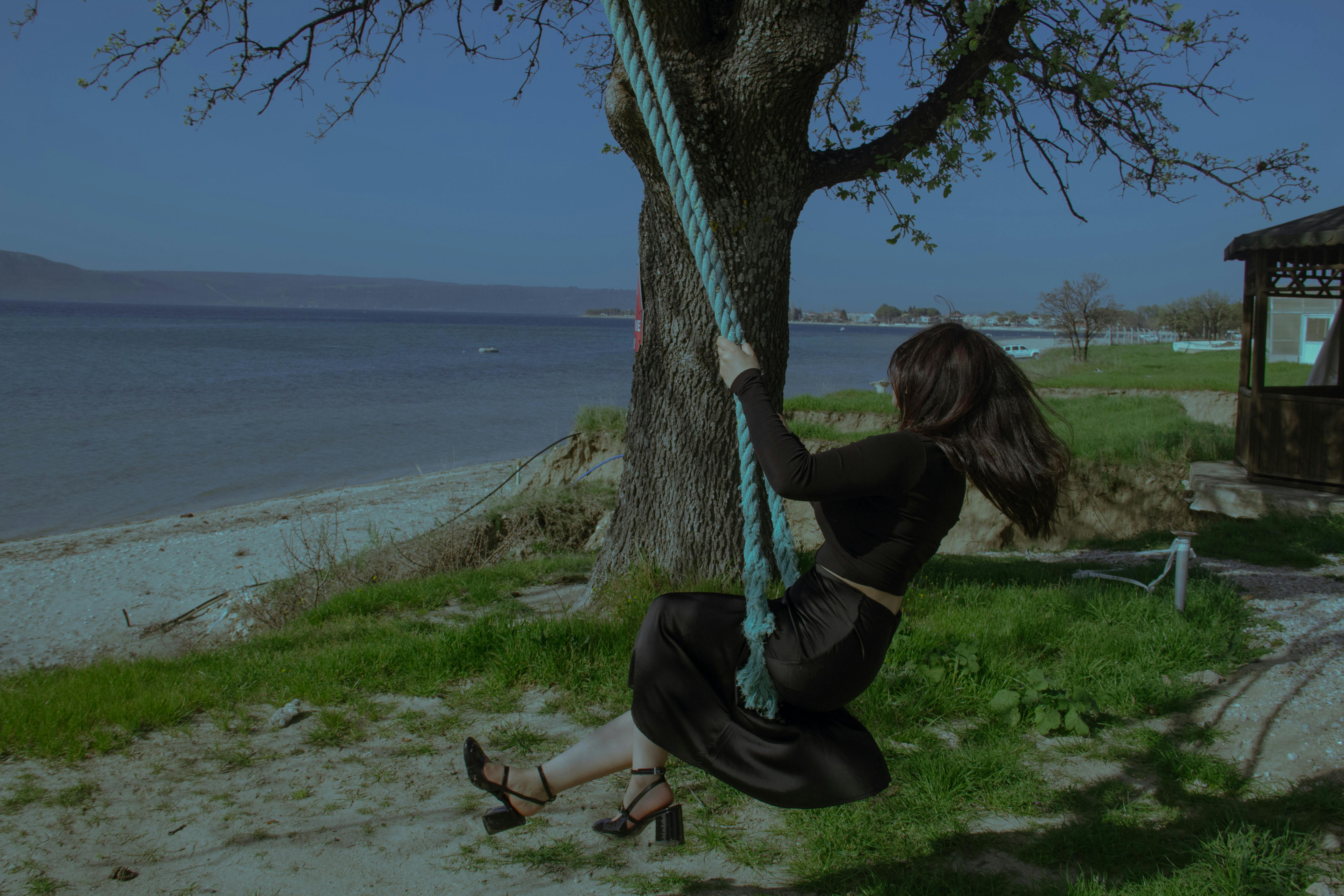 A woman in a black outfit enjoys swinging on a rope swing by a serene lakeshore.