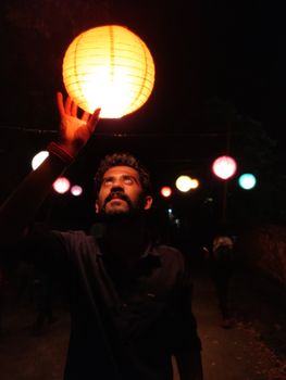 Man holding a glowing lantern at a nighttime festival, surrounded by colorful hanging lights.