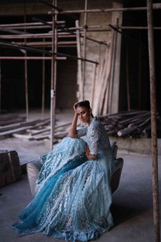 Woman in a shimmering blue dress seated in an unfinished indoor construction site, conveying elegance.