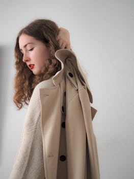 Portrait of a woman with long brown hair holding a beige coat against a white background.