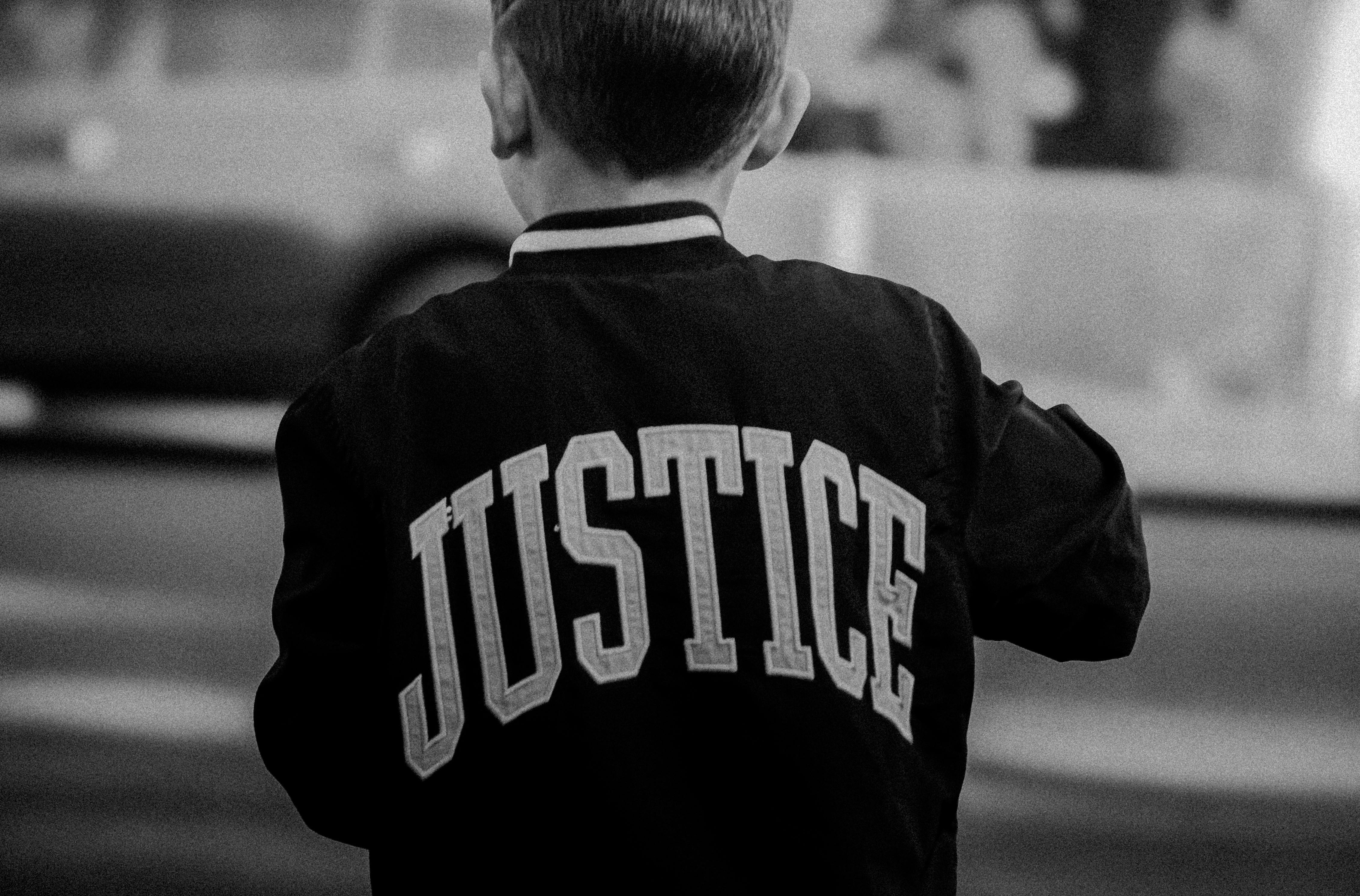 A child standing in an urban area wearing a jacket with the word 'Justice' printed on the back.