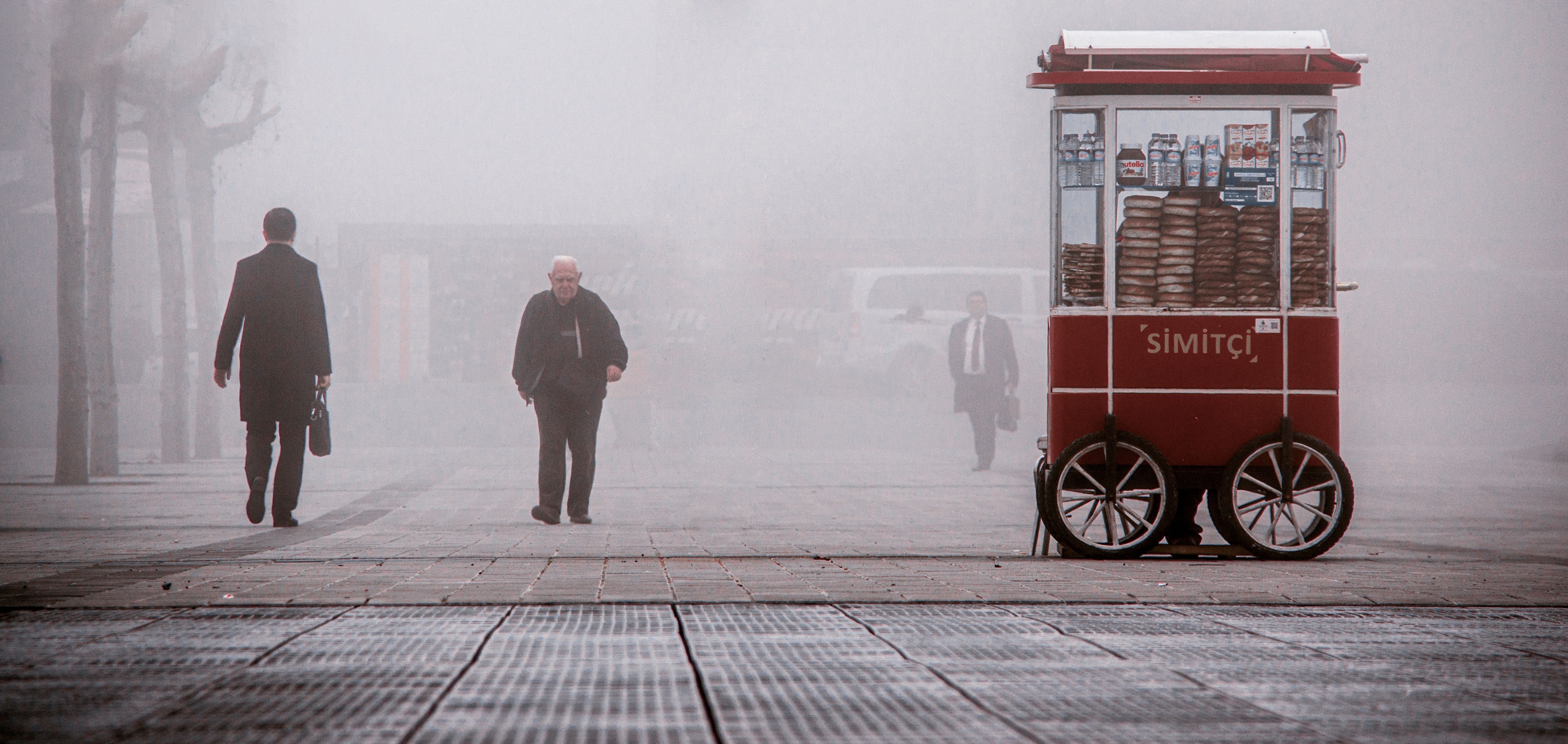 A foggy urban scene featuring a simit cart and pedestrians walking in a misty square, creating a moody atmosphere.