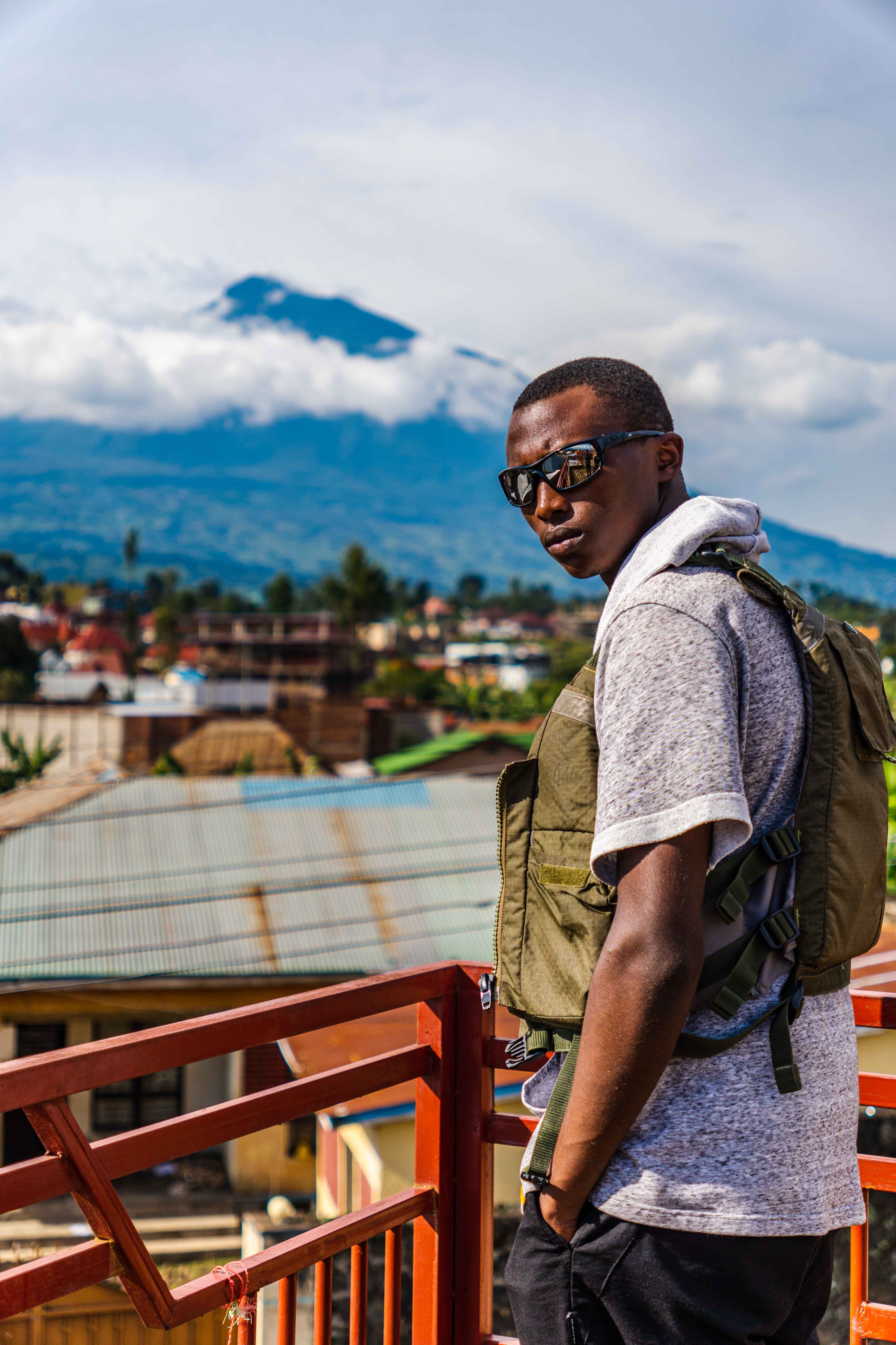 Man in vest poses on a balcony with a mountain view, cityscape and vibrant sky.