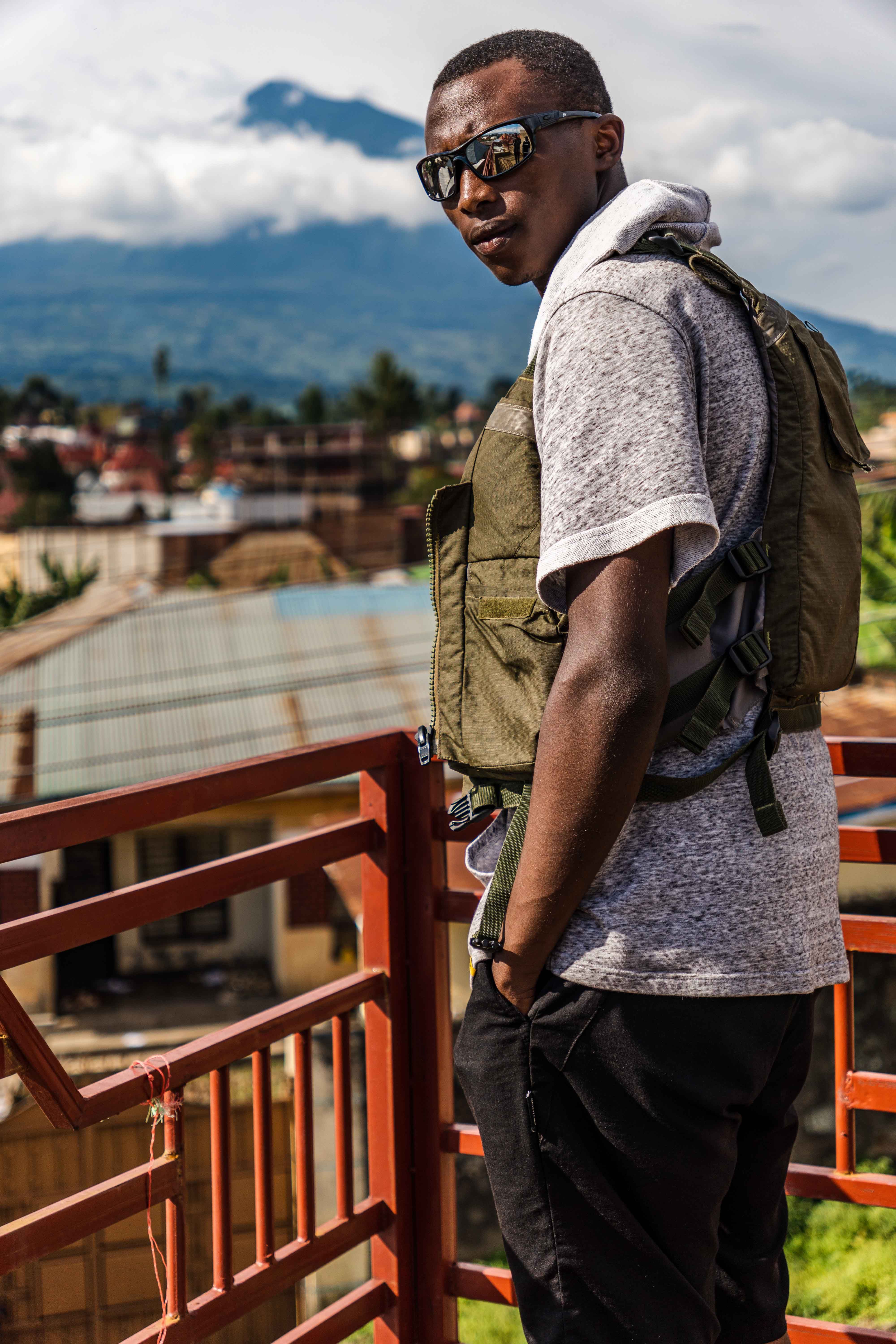 Stylishly dressed man in sunglasses stands on a rooftop overlooking a cityscape with a mountainous backdrop.