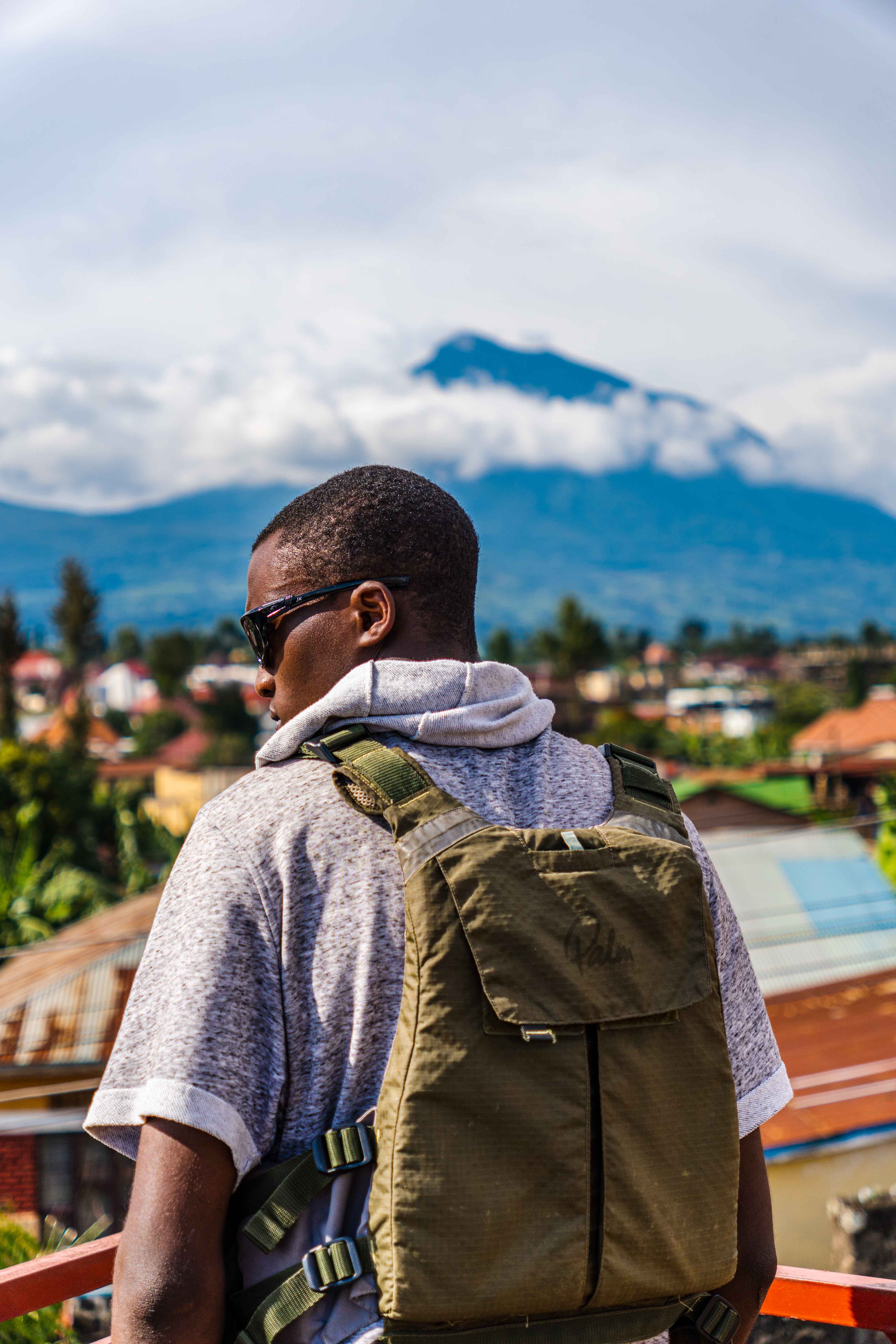 A man wearing sunglasses and a backpack stands on a balcony, gazing at a distant mountain with an urban background.