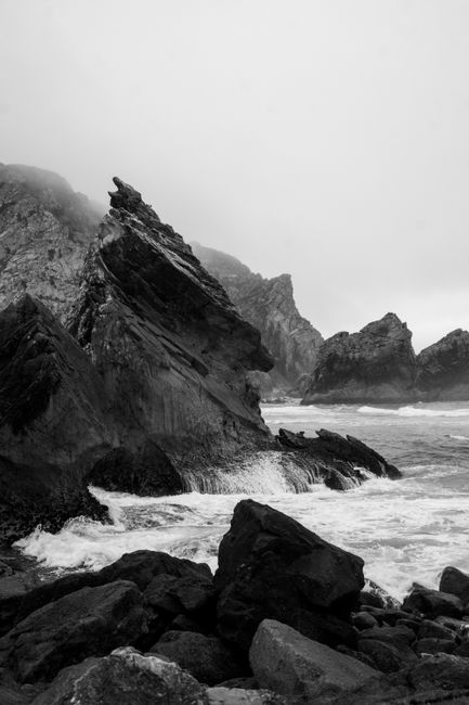 Monochrome photograph showcasing rugged coastal rocks with crashing waves, conveying a dramatic and natural vibe.