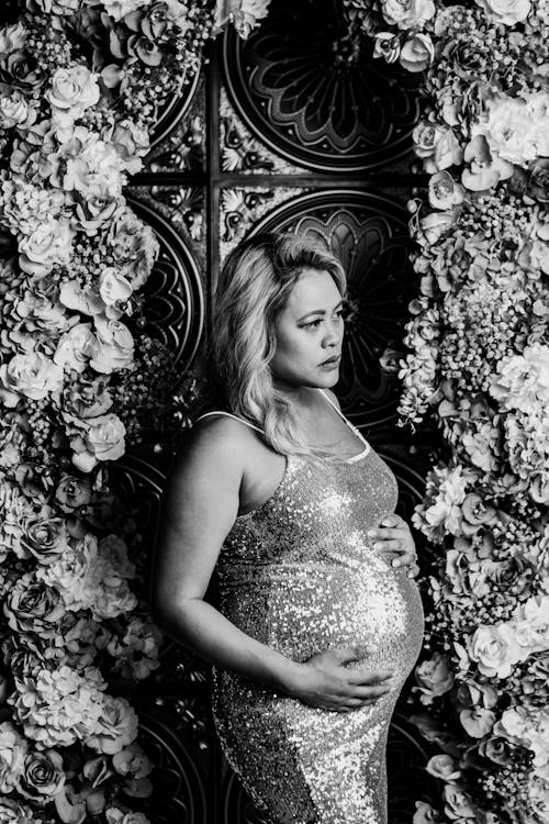 A pregnant woman in a silver dress is standing in front of a floral wall
