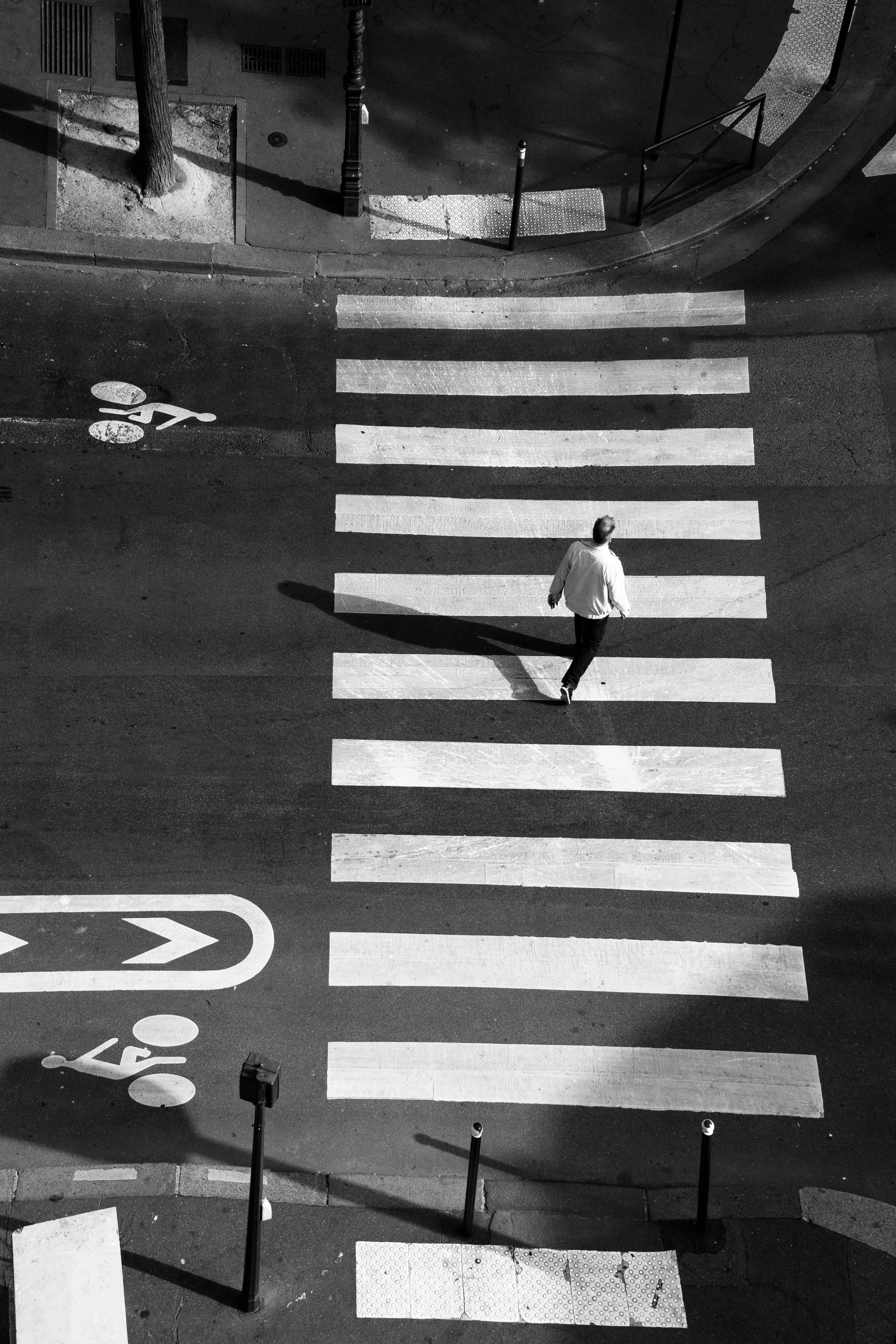 A person crossing an urban street at a crosswalk, photographed from above in black and white.